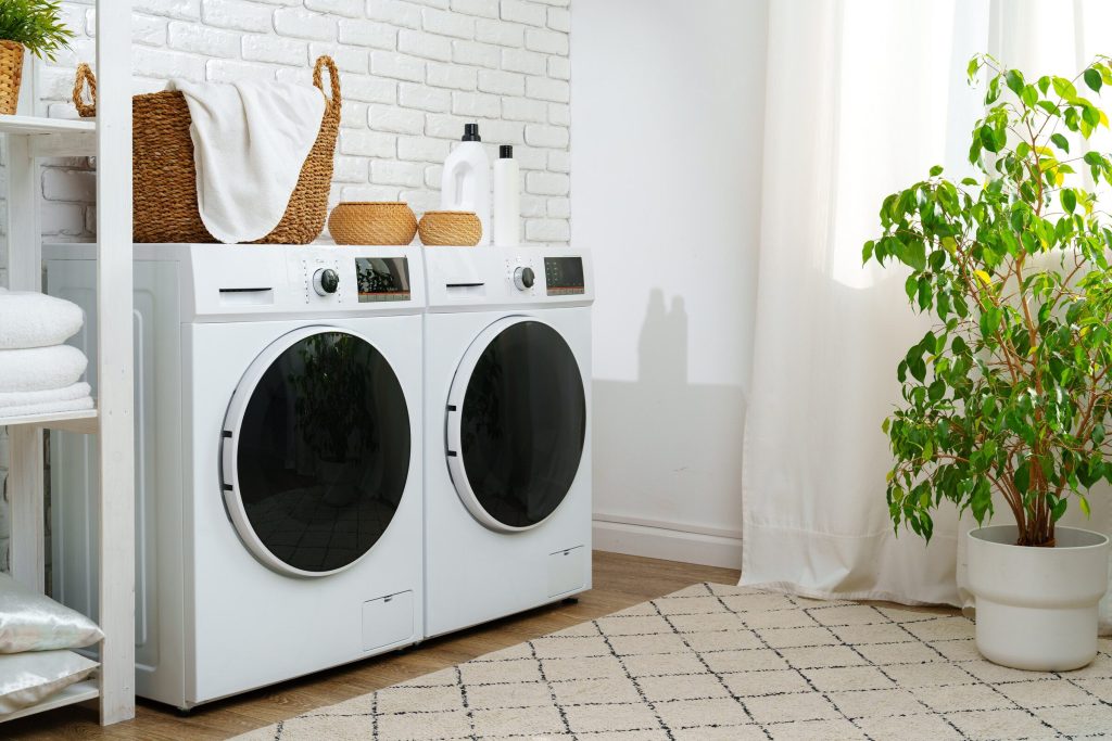 Two white front-loading washing machines side by side in a bright laundry room with a white brick wall