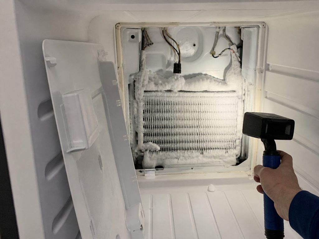 The rear panel of a freezer compartment removed and leaning aside, the evaporator coil behind it thick with white frost, a work light across it.