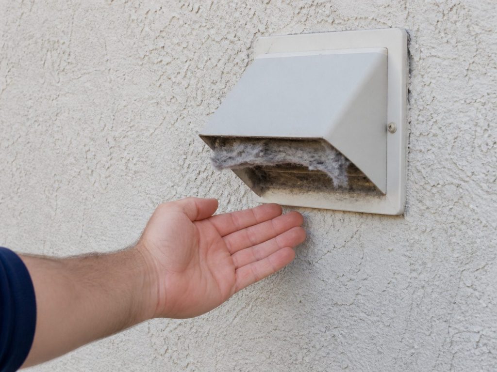 The outside wall of an American house with a dryer vent hood, its flap partly propped open by grey lint, a hand held in front of it feeling for airflow.