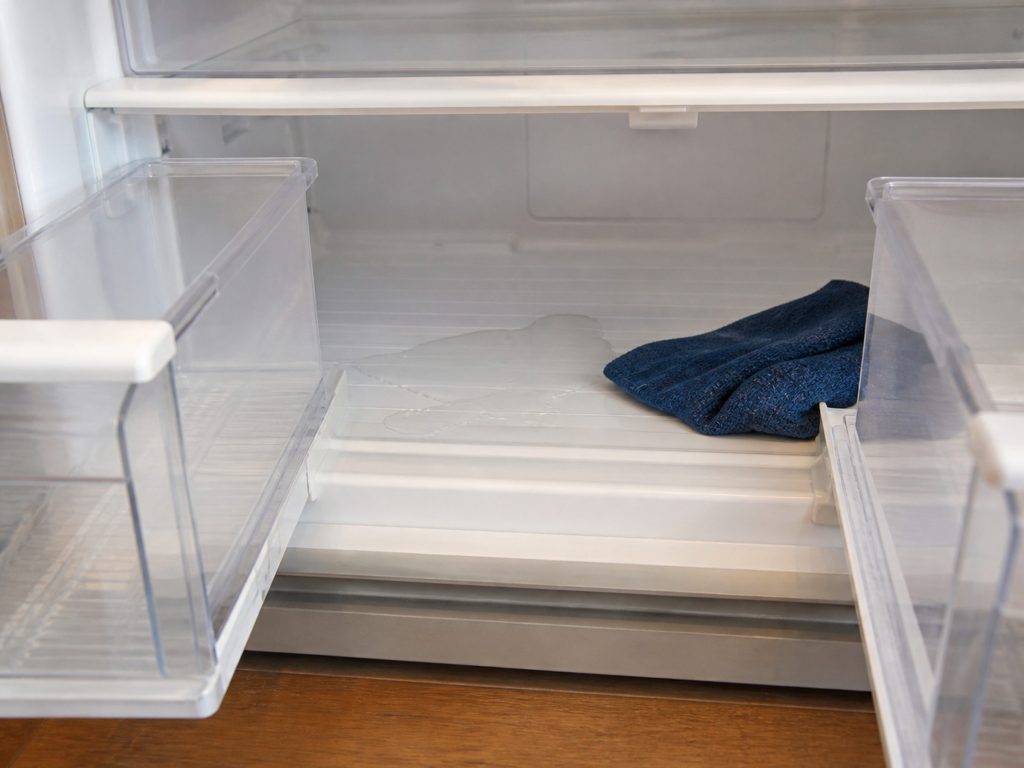 The bottom of a refrigerator interior with the crisper drawers pulled out and a shallow pool of water lying on the liner beneath them, a cloth beside it.