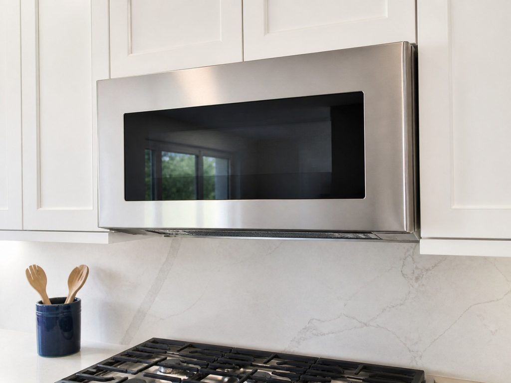 A stainless steel over-the-range microwave mounted above a cooktop in a bright white American kitchen, door closed, plain unmarked fascia, no badge and no lettering, the vent grille visible along its underside.